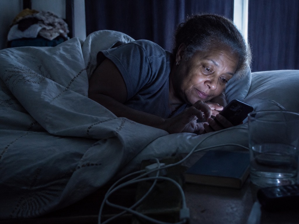 An older Black woman with greying hair lies on her side in bed in a dim room, her face lit by the cool blue glow of the smartphone she is scrolling, with a tangled white charger cable, a closed hardcover book and a half-full water glass on the nightstand.